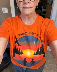 A close-up selfie of a woman wearing an orange t-shirt with a colorful sunset design and the phrase 'Every Child Matters.' The design features two silhouettes holding hands in front of a forest and a starry sky.