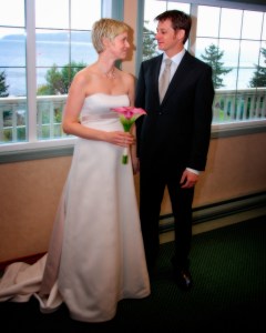A bride in a strapless white wedding gown holding a pink flower bouquet, standing with a groom in a black suit, both looking at each other with a scenic view behind them.